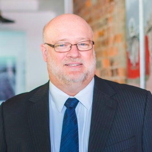 Photo of Simon Levers upper torso.  He is wearing a jacket and tie and has seeing glasses on.