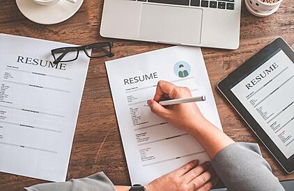 Photo of paper work, with resume at the top and a computer and reading glasses off to the side. a persons hand are over the paperwork with a pen in the right hand.