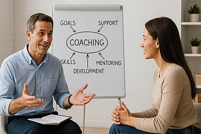 Two people sitting and talking with one gesturing with  their hands.  A white board in the background says "coaching"in the middle with the words, "goals, support, mentoring, development and skills" around the outside.