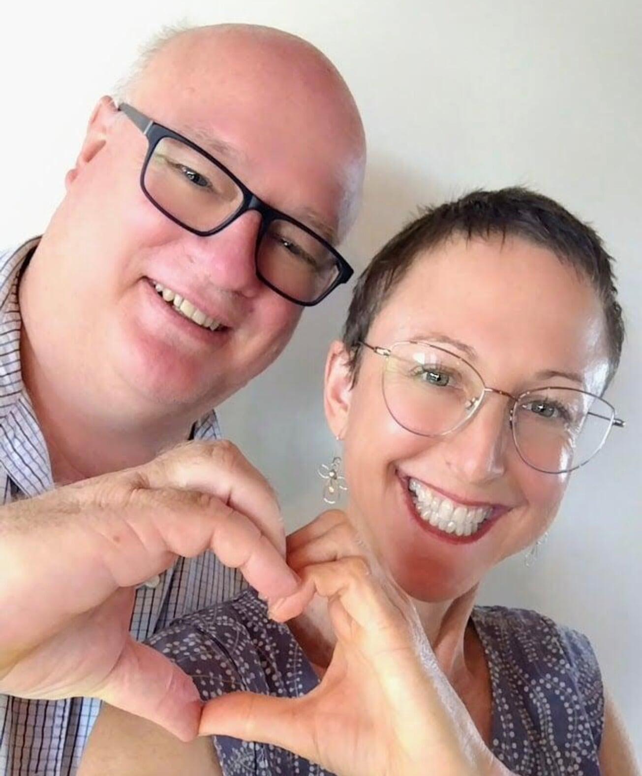 Photo of Simon and Brooke just their heads and their hands that are joining together to make a love heart shape.  Both are wearing reading glasses,